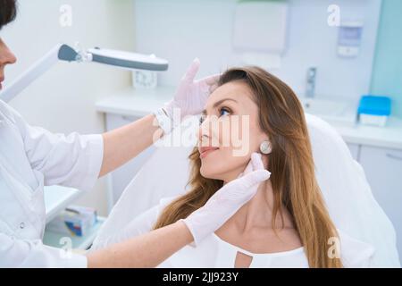 Medico che esamina un volto di donna con entrambe le mani gloved Foto Stock