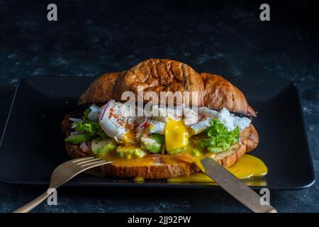 Croissant sandwich fresco con uova in camicia, salmone e avocado su un piatto su sfondo nero, primo piano Foto Stock