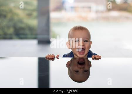 Ride carino naughty bambino ritratto con la testa sopra il tavolo di vetro con riflessione, bambino positivo casa vita concetto scena Foto Stock