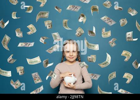 Ragazza piccola felice con bianco Piggy banca in dollari americani pioggia di denaro su sfondo blu Foto Stock
