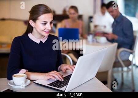 sorridente giovane donna che usa il computer portatile e beve caffè in un bar Foto Stock
