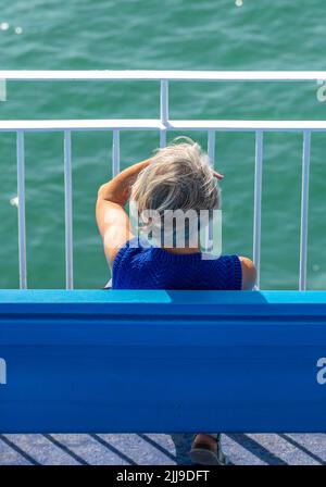 signora anziana seduta da sola su una panchina che guarda fuori al mare, femmina matura nel profondo del pensiero, solitudine, signora seduta da sola ombreggiando i suoi occhi dal sole Foto Stock