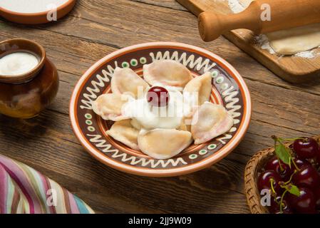 Gnocchi ucraini tradizionali, vareniki con ciliegie in una ciotola dipinta in ceramica con ingredienti su un vecchio tavolo rurale in legno. Foto Stock