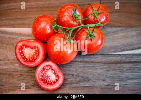 Pomodori rossi su vite in cucina - pomodori freschi a fette e integrali su tagliere di legno - grappolo e grappolo Solanum lycopersicum maturi Foto Stock