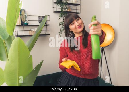 Donna caucasica sorridente con capelli lunghi e scuri che tiene gli attrezzi di pulizia in salotto vicino alla pianta guardando la macchina fotografica. Scatto al coperto. Foto di alta qualità Foto Stock