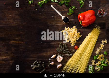 Vari tipi di pasta su sfondo di legno scuro. FARFALLE, fusilli, spaghetti, peperone, insalata di mais, spicchi d'aglio rossi, verdi o gialli Foto Stock