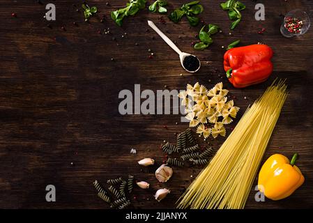 Vari tipi di pasta su sfondo di legno scuro. FARFALLE, fusilli, spaghetti, peperone, insalata di mais, spicchi d'aglio rossi, verdi o gialli Foto Stock