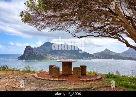 La panca pubblica si affaccia sull'oceano e la montagna sembra bella fuori nella natura. Paesaggio di quattro sedie vuote e un tavolo con un albero che le dà Foto Stock
