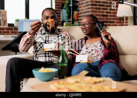 Uomo che mangia tagliatelle da asporto con bacchette e donna che usa il telecomando per cambiare canale tv. Goditi un pasto con consegna da asporto insieme e guarda un film in programma in televisione. Foto Stock