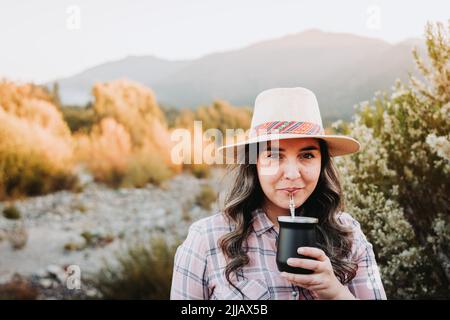 Donna sorridente con un cappello e indossa una blusa rosa pallido, bevendo il compagno in uno spazio naturale al tramonto Foto Stock