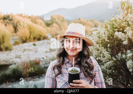 Donna sorridente con un cappello e indossa una blusa rosa pallido, bevendo il compagno in uno spazio naturale al tramonto Foto Stock