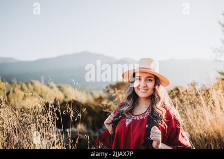 Donna zaino in spalla latino-americana che indossa un poncho rosso tradizionale e un cappello in uno spazio naturale soleggiato Foto Stock