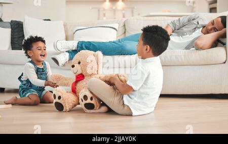 Il papà ha bisogno di una pausa: Due fratelli che combattono su un teddy sul pavimento mentre il loro papà dorme sul divano a casa. Foto Stock