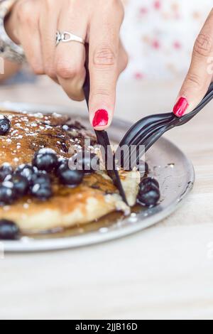 Mani che usano argenteria di plastica per tagliare i pancake di mirtillo e blackberry Foto Stock