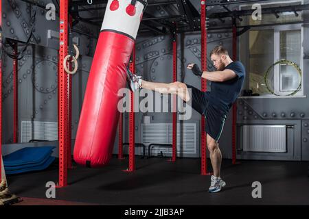 Giovani forti bearded arti marziali miste combattenti treni in palestra. Offre un potente calcio dritto al sacchetto da punzonatura rosso. Foto Stock