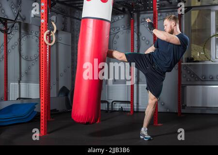 Giovani forti bearded arti marziali miste combattenti treni in palestra. Offre un potente calcio laterale al sacchetto per punzonatura rosso. Foto Stock