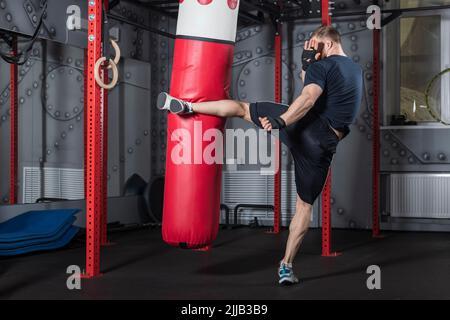 Young strong bearded mixed martial arts fighter trains in gym. Delivers powerful side kick to red punching bag. Stock Photo