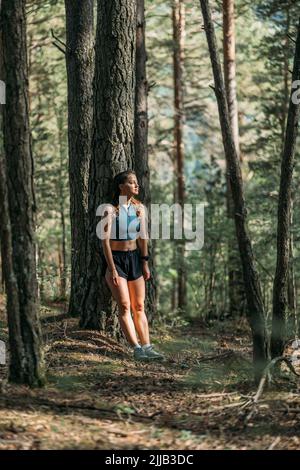 Giovane donna stanca in abbigliamento sportivo appoggiata su un albero e riposata nella foresta dopo essersi allenata. Trail running e concetto di vita attiva. Foto Stock