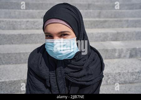 Donna in un hijab siede sulle scale indossando una maschera Foto Stock