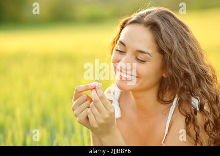 Donna felice guardando anello di impegno seduto in un campo di grano Foto Stock