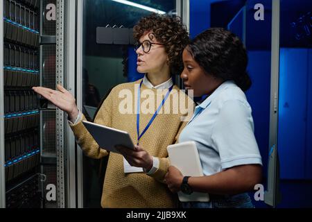 Due ingegneri femminili che utilizzano un tablet pc nel loro lavoro di squadra, si trovano vicino al rack server aperto e discutono della connessione nel data center Foto Stock