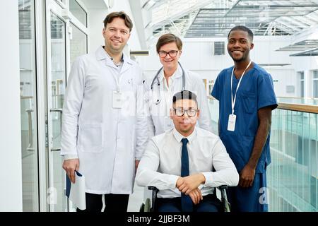 Gruppo di medici diversi che posano con il paziente in sedia a rotelle presso la clinica moderna, tutti sorridenti alla macchina fotografica Foto Stock