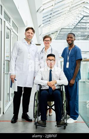 Ritratto a tutta lunghezza diversi gruppi di medici in posa con paziente in sedia a rotelle in clinica moderna, tutti guardando la macchina fotografica e sorridente Foto Stock