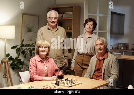 Due allegre coppie anziane in casualwear in piedi e seduti al tavolo con scacchiera e bicchieri di vino rosso mentre vi godete la festa di casa Foto Stock