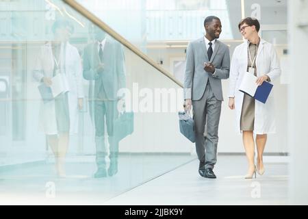 Ritratto minimo a tutta lunghezza di uomo d'affari nero sorridente che parla con collega femmina mentre cammina verso la macchina fotografica nella sala dell'edificio d'ufficio, copia sp Foto Stock