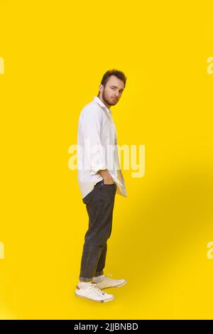 Giovane uomo pensivo e premuroso in camicia bianca e jeans denim che si posa su sfondo giallo con le mani in tasca. Elegante uomo hipster dal look casual elegante. Foto Stock