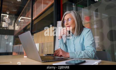 Donna d'affari anziana seduta sul posto di lavoro e guardando la macchina fotografica Foto Stock