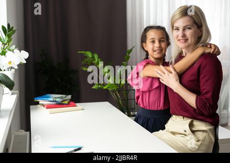 Madre che si prende cura calmando e abbracciando la figlia piccola, seduta alla scrivania insieme, mamma che ama esprimere il sostegno, confortante offesa ragazza prescolare Foto Stock