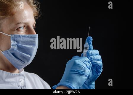 Una donna che indossa una maschera protettiva e guanti tiene una siringa con un vaccino o un'iniezione su sfondo nero. Si prepara un'infermiera femminile Foto Stock