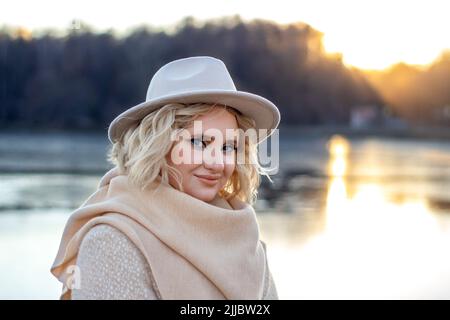 Ritratto di una donna bionda con un misterioso sorriso modesto in un cappello e stole con occhio luminoso trucco in autunno nei raggi del sole che tramonta contro t Foto Stock