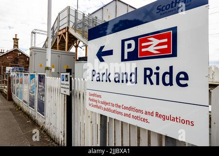 Primo piano di un cartello Park and Ride alla stazione ferroviaria di Cardross ScotRail, con un cartello a P e una freccia per il parcheggio, con una recinzione bianca per picchetti e un ponte pedonale Foto Stock