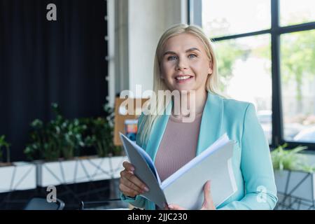 Psicologo sorridente che tiene la cartella della carta e guarda la macchina fotografica nella sala di consultazione Foto Stock