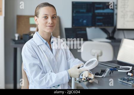 Ritratto di giovane scienziata femminile sorridente alla macchina fotografica mentre si siede sul posto di lavoro in laboratorio di ingegneria, spazio copia Foto Stock