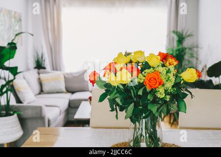 Tavolo da cucina con fuoco su vaso con rose gialle e arancioni su di esso con sfondo sfocato di moderno e accogliente soggiorno con divano e verde pl Foto Stock