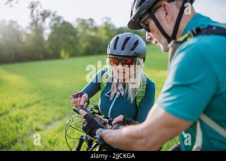 Coppia attiva di anziani che riposano dopo un giro in bicicletta nel parco estivo, utilizzando lo smartphone. Foto Stock