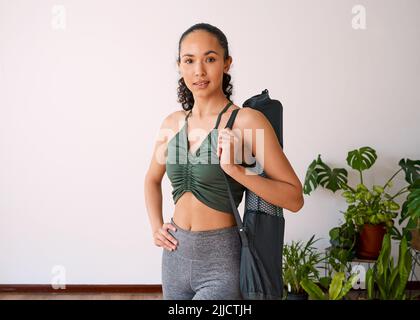 Una giovane donna multietnica si alza con il suo tappetino yoga in studio pieno di piante Foto Stock