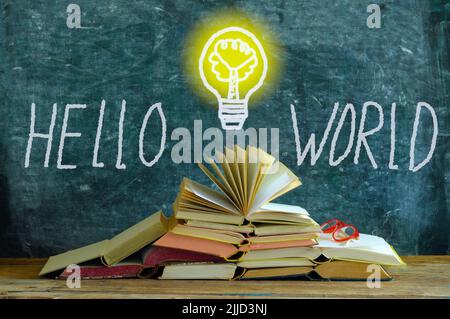 pila di libri di fronte a una lavagna, disegno di una lampadina e slogan hello mondo, conoscenza, istruzione e concetto di integrazione sociale Foto Stock
