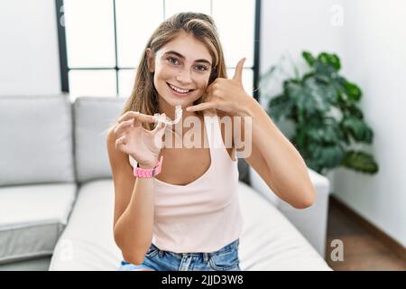 Giovane donna bionda che tiene invisibile allineatore ortodontico sorridendo facendo il gesto del telefono con la mano e le dita come parlare al telefono. Comunicatin Foto Stock