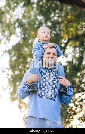 Felice padre porta suo figlio sulle spalle durante la passeggiata sul parco. Sono vestiti con camicie ricamate nazionali ucraine. Foto Stock