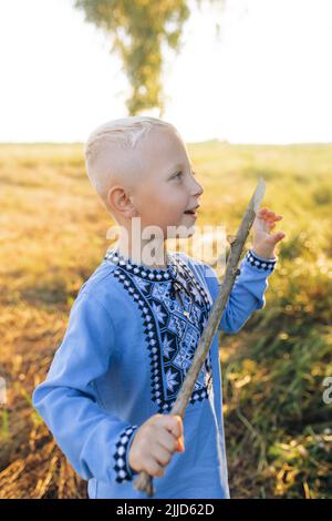 Il bambino felice ha divertimento e gioca sul prato con bastone di legno. È vestito in maglia nazionale ricamata Ucraina. Foto Stock