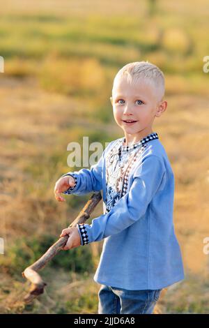 Il bambino felice ha divertimento e gioca sul prato con bastone di legno. È vestito in maglia nazionale ricamata Ucraina. Foto Stock