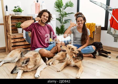 Giovane coppia ispanica che fa la lavanderia con i cani che sorridono facendo il gesto del telefono con la mano e le dita come parlare al telefono. Comunicare i concetti. Foto Stock