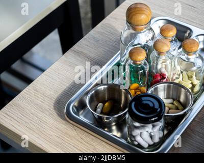 Primo piano set di vitamine e integratori nelle piccole bottiglie di vetro trasparente nel vassoio medico in acciaio su tavola di legno. Vitamina verde, gialla, rossa, bianca Foto Stock