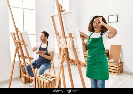Ispanica donna di mezza età e uomo maturo in studio d'arte sorridendo felice di fare ok segno con mano su occhio guardando attraverso le dita Foto Stock