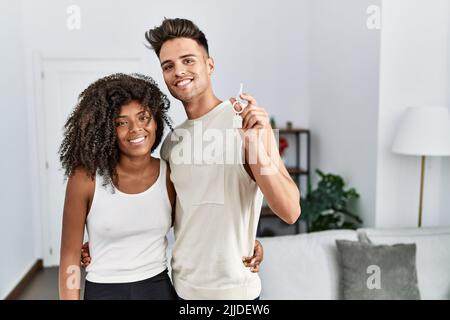 Giovane coppia interrazziale che tiene le chiavi della nuova casa guardando positivo e felice in piedi e sorridendo con un sorriso sicuro che mostra i denti Foto Stock