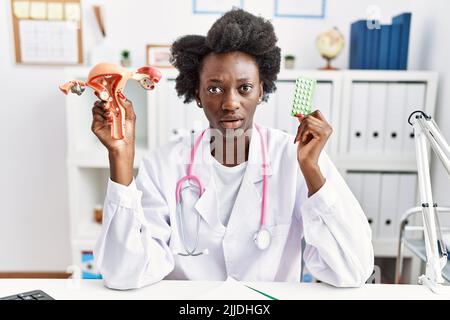 Medico africano donna che tiene anatomico organo genitale femminile e pillole di controllo delle nascite clueless e confuso espressione. Concetto di dubbio. Foto Stock